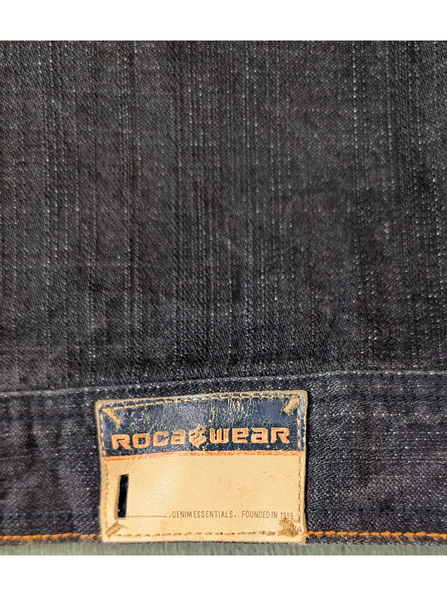 JAKE & IPI Rocawear dark wash denim jacket with leather logo patch and "DENIM ESSENTIALS. FOUNDED IN 1999" text.