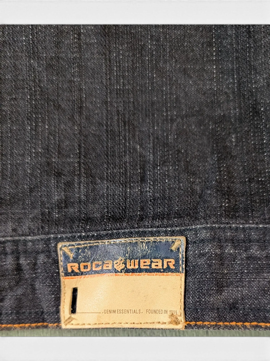 JAKE & IPI Rocawear dark wash denim jacket with leather logo patch and "DENIM ESSENTIALS. FOUNDED IN 1999" text.
