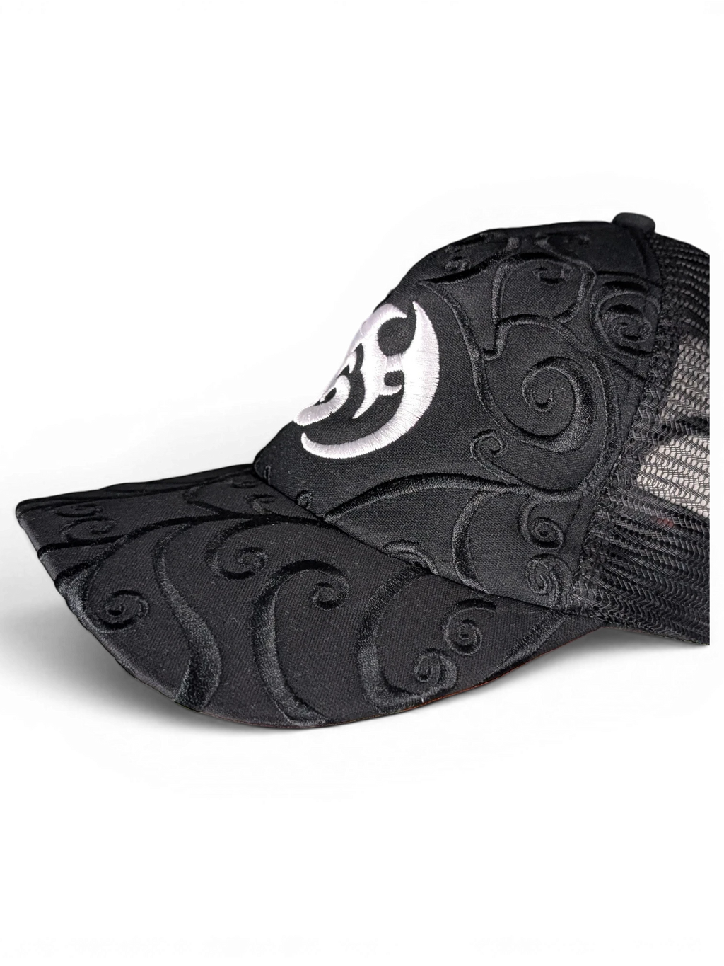 JAKE & IPI black trucker cap featuring intricate tonal swirl embroidery and a white circular logo on the front panel.