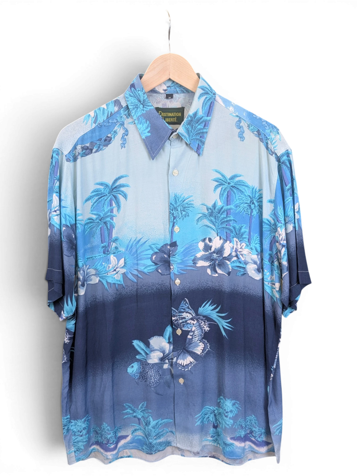 JAKE & IPI Destination Liberté blue Hawaiian shirt with tropical palm tree and floral print on a wooden hanger