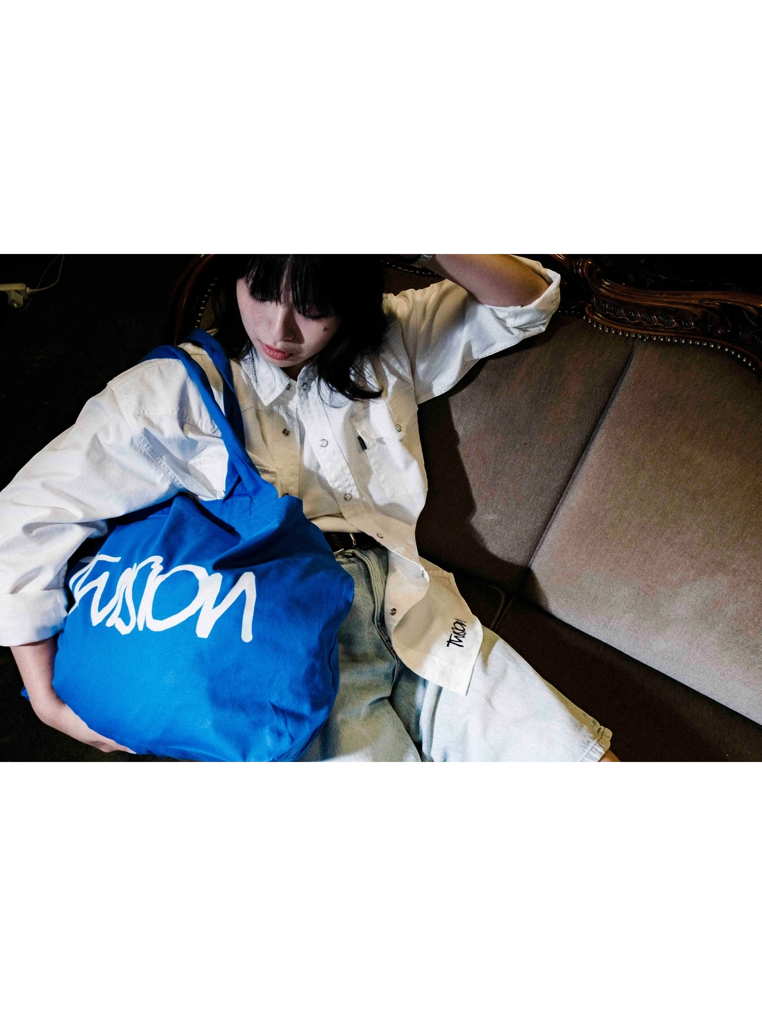A person wearing a JAKE & IPI white Twision shirt and light wash jeans, holding a bright blue tote bag.