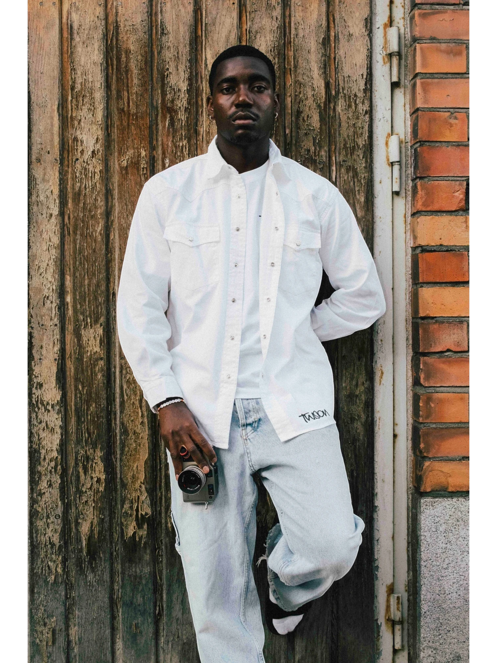 A man wearing a white JAKE & IPI Twision shirt over a white t-shirt and light wash jeans, leaning against a door.