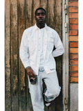 A man wearing a white JAKE & IPI Twision shirt over a white t-shirt and light wash jeans, leaning against a door.
