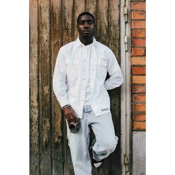 A man wearing a white JAKE & IPI Twision shirt over a white t-shirt and light wash jeans, leaning against a door.