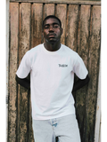 A man wearing a white JAKE & IPI Twision T-shirt with a black logo graphic, standing against a rustic wooden door.