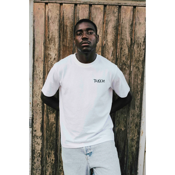 A man wearing a white JAKE & IPI Twision T-shirt with a black logo graphic, standing against a rustic wooden door.