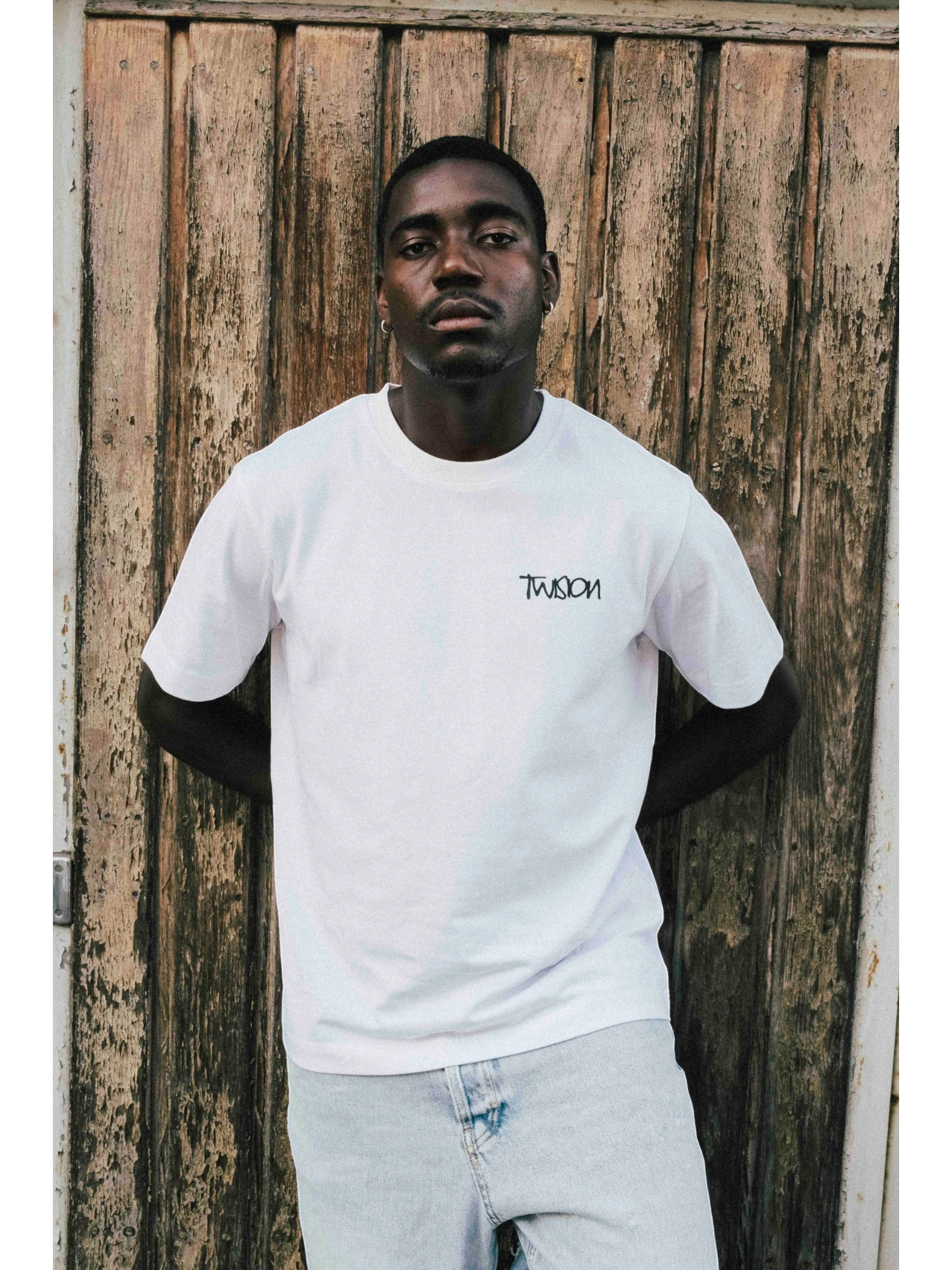 A man wearing a white JAKE & IPI Twision T-shirt with a black logo graphic, standing against a rustic wooden door.