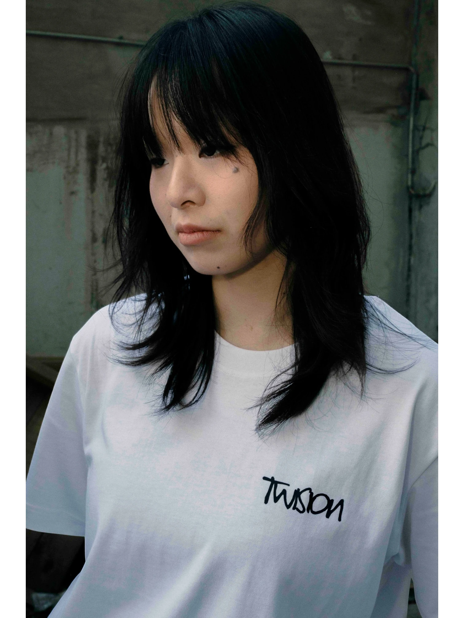 A woman wearing a white JAKE & IPI Twision T-shirt with black script logo on the chest against a concrete background.